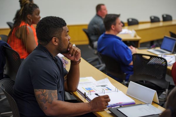 Man in black t-shirt listens and takes notes during a lecture. 