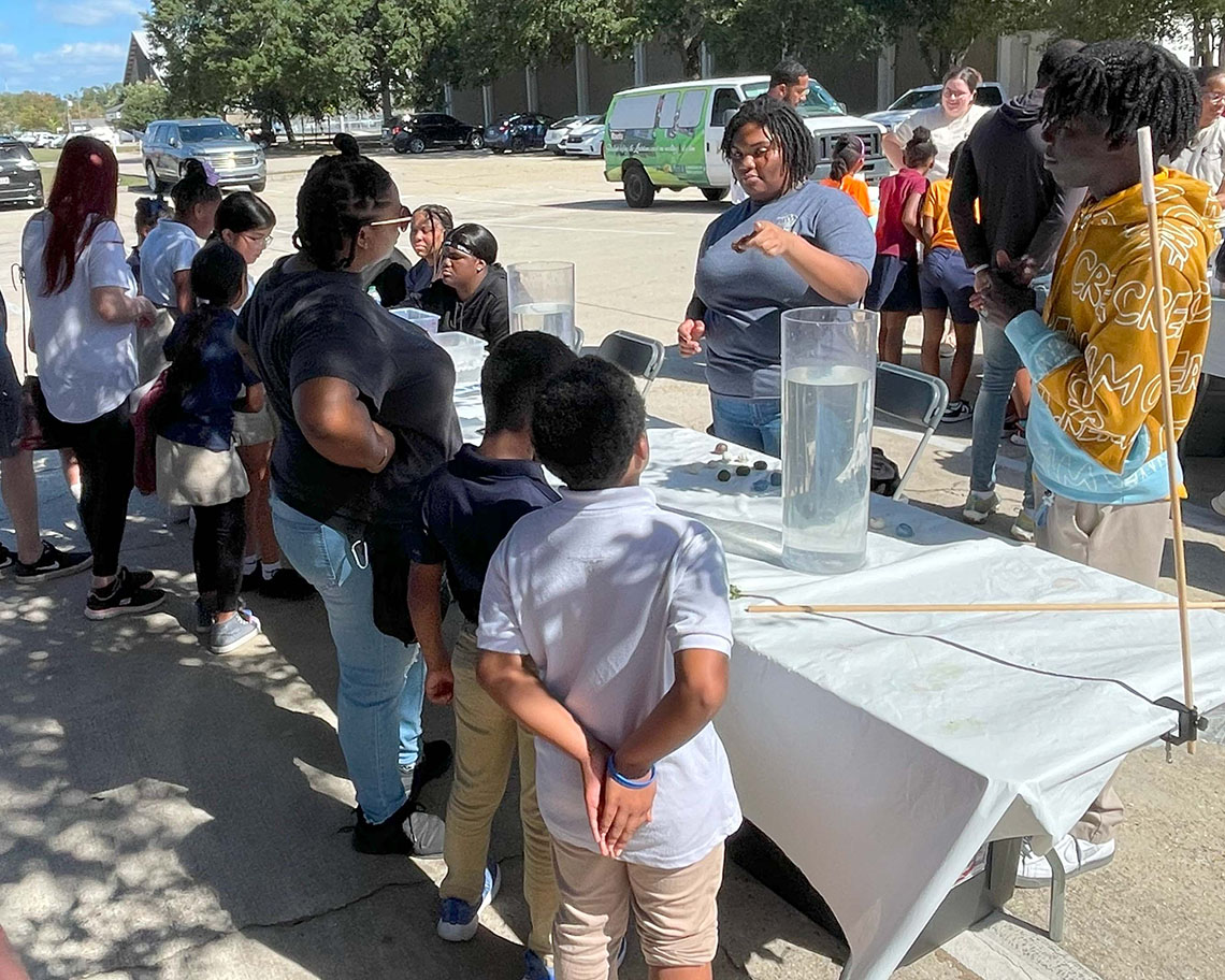 two students stand behind a table talking to a group of children